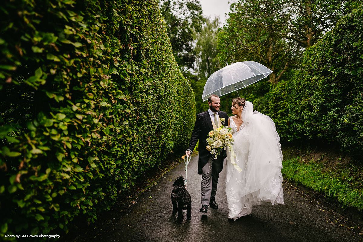 Bride and groom with their dog, walking in the gardens of Hilltop Country House carrying a see-through umbrella.