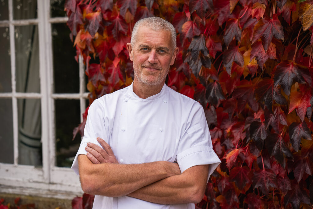 Featuring Chef Paul Merrett, this image showcases him in a serene outdoor setting, underscored by the vivid autumnal colors of the foliage. His pose, relaxed yet confident, and his direct gaze convey both his experience and approachability. Dressed in a classic white chef’s jacket, he stands as a symbol of culinary expertise and creativity. The backdrop, rich with the hues of fall, subtly hints at the natural, seasonal ingredients that likely inspire his dishes. This portrait not only highlights Chef Merrett’s professional stature but also his connection to the natural world, suggesting a cooking philosophy that values freshness, seasonality, and the beauty of the environment.