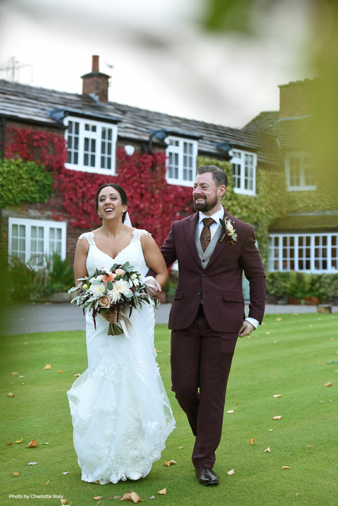 A joyous bride and groom stroll hand in hand on the manicured lawns of Hilltop, the bride in a white lace gown with a bouquet of autumnal flowers, and the groom in a stylish burgundy suit that echoes the warm, on-trend colors of the season, set against a classic country house adorned with vibrant red ivy.