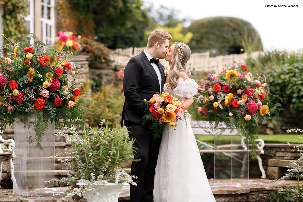 A bride and groom sharing a kiss at an elegant garden wedding venue. The bride holds a vibrant bouquet of orange, yellow, and pink flowers, complementing her off-the-shoulder wedding gown, while the groom dons a classic black tuxedo. They are surrounded by lush floral arrangements and greenery, with the serene ambience of Hilltop Country House, creating a picturesque setting for a seasonal wedding theme.