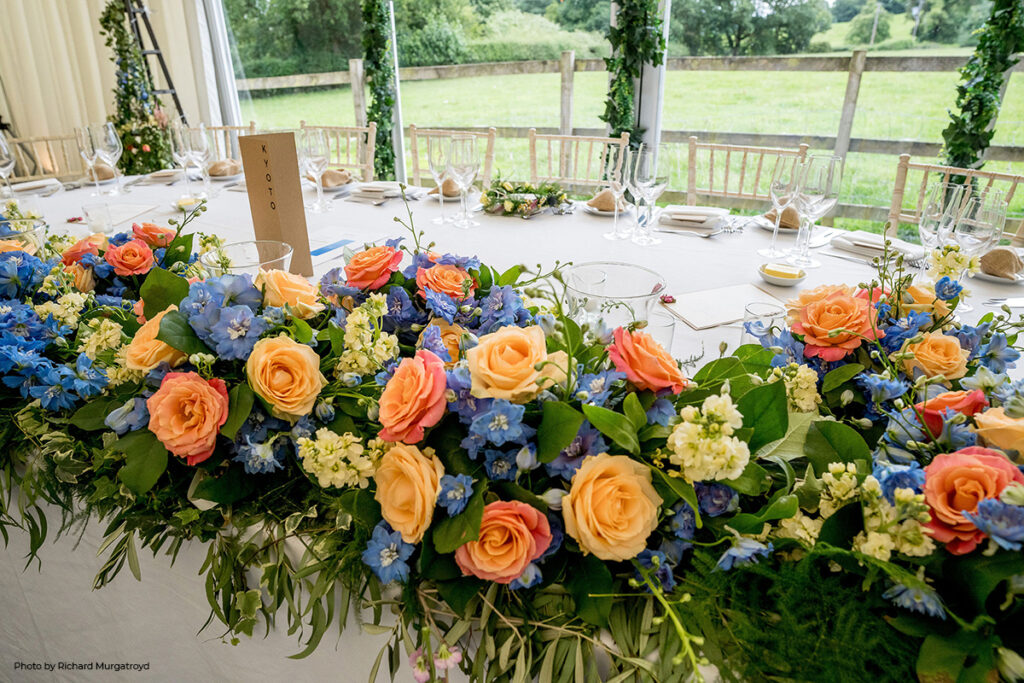 A lush wedding table centerpiece brimming with spring colors: vibrant orange roses and bluebells are nestled among a variety of seasonal greenery, creating a striking contrast that perfectly captures the essence of spring at Hilltop.