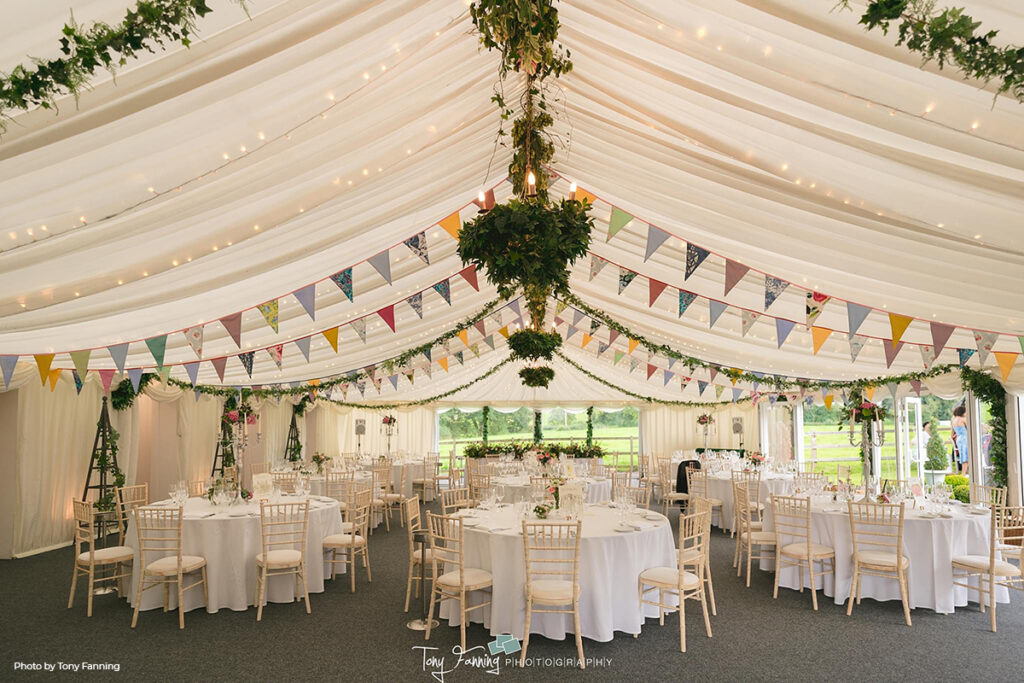 nterior view of a marquee at Hilltop Country House, elegantly set up for a wedding reception with white tables and chairs, beautifully complemented by colorful bunting draped across the ceiling, enhancing the summertime garden celebration theme