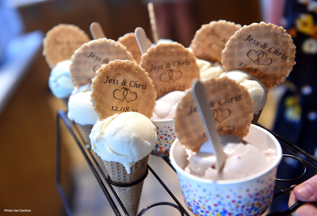 Close-up of personalized ice cream wafers, each inscribed with 'Jess & Chris 12.08.23', stuck in scoops of ice cream in various cones, offering a bespoke touch to a summer wedding treat