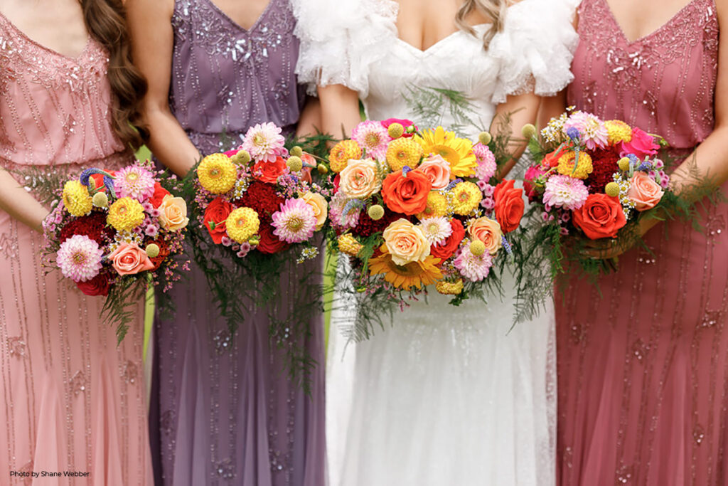 A row of bridesmaids in pastel pink and purple gowns, holding vibrant wedding bouquets with an array of summer flowers in pink, yellow, red, and orange, embodying the colorful essence of a summer wedding at Hilltop.