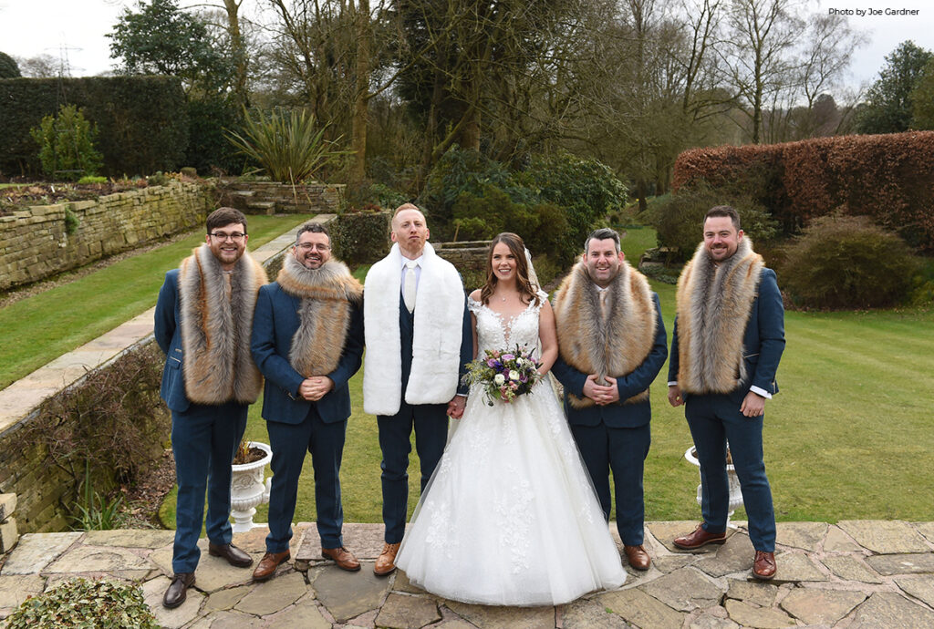 A bridal party stands outdoors at Hilltop, the men wearing stylish navy suits paired with luxurious fur shawls, embracing the chill of a winter wedding, while the bride in her elegant gown and holding a bouquet adds a touch of timeless grace.