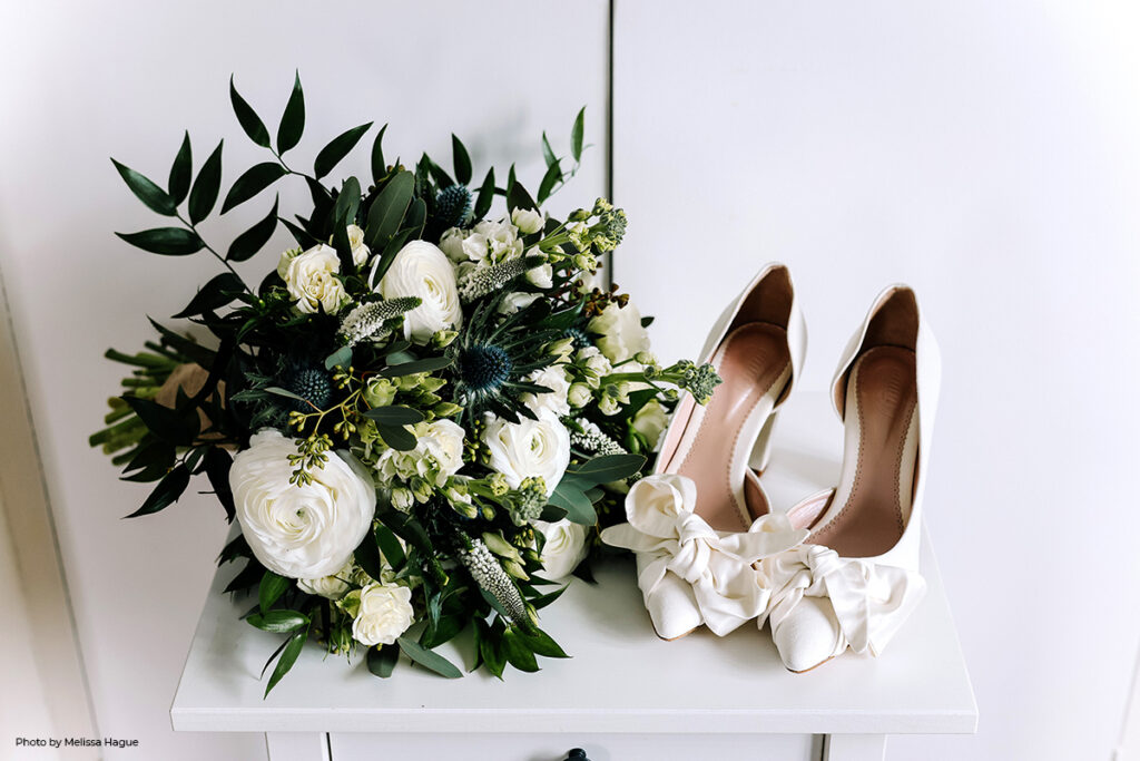 An elegant bridal bouquet featuring white roses and green foliage, paired with chic bridal shoes adorned with a cream ribbon, captures the classic winter wedding color palette of sage, emerald green, and creams.